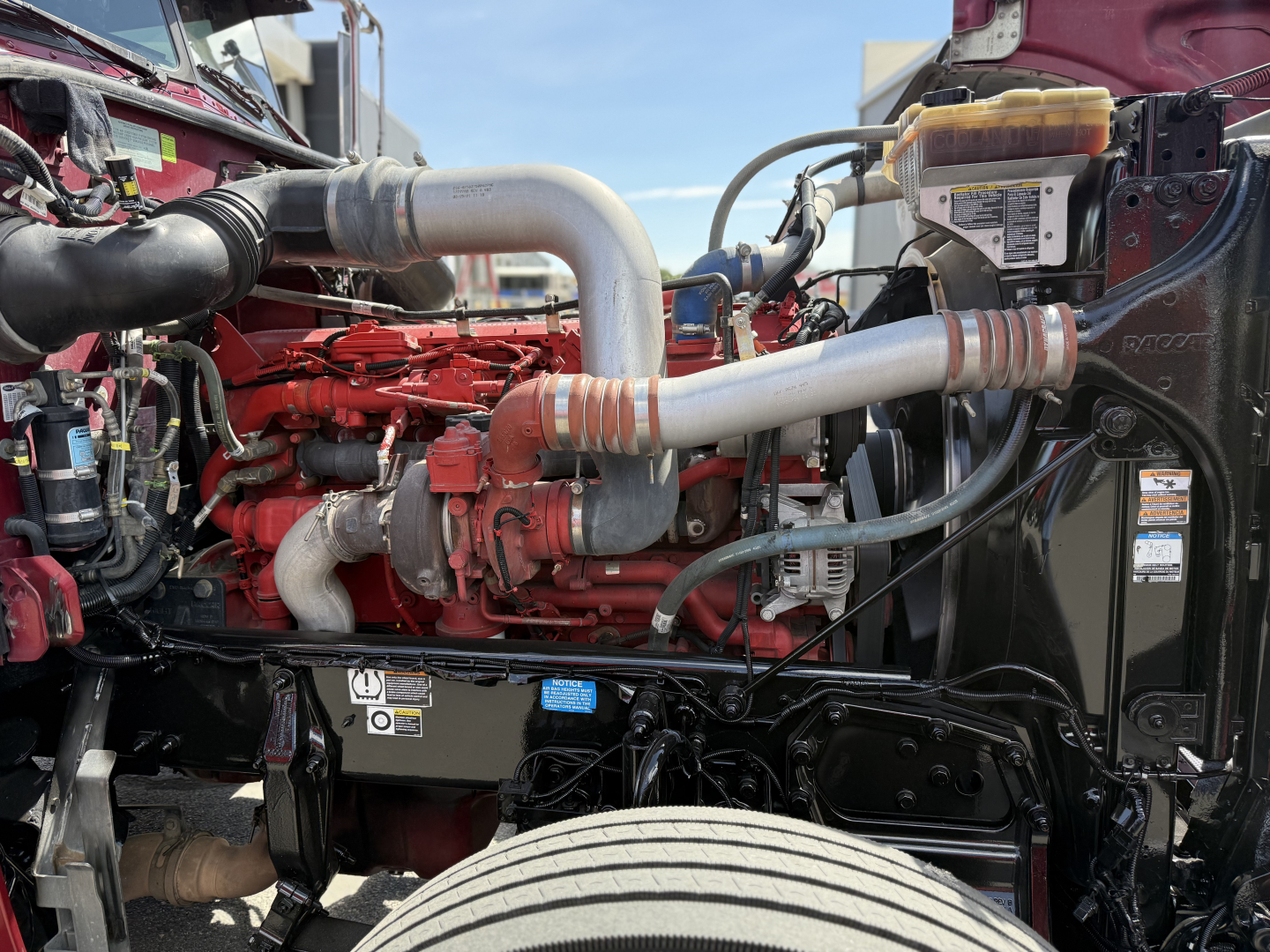Peterbilt 389 engine bay showing red diesel engine with intake manifold, silver exhaust piping, coolant reservoir, and various hoses.