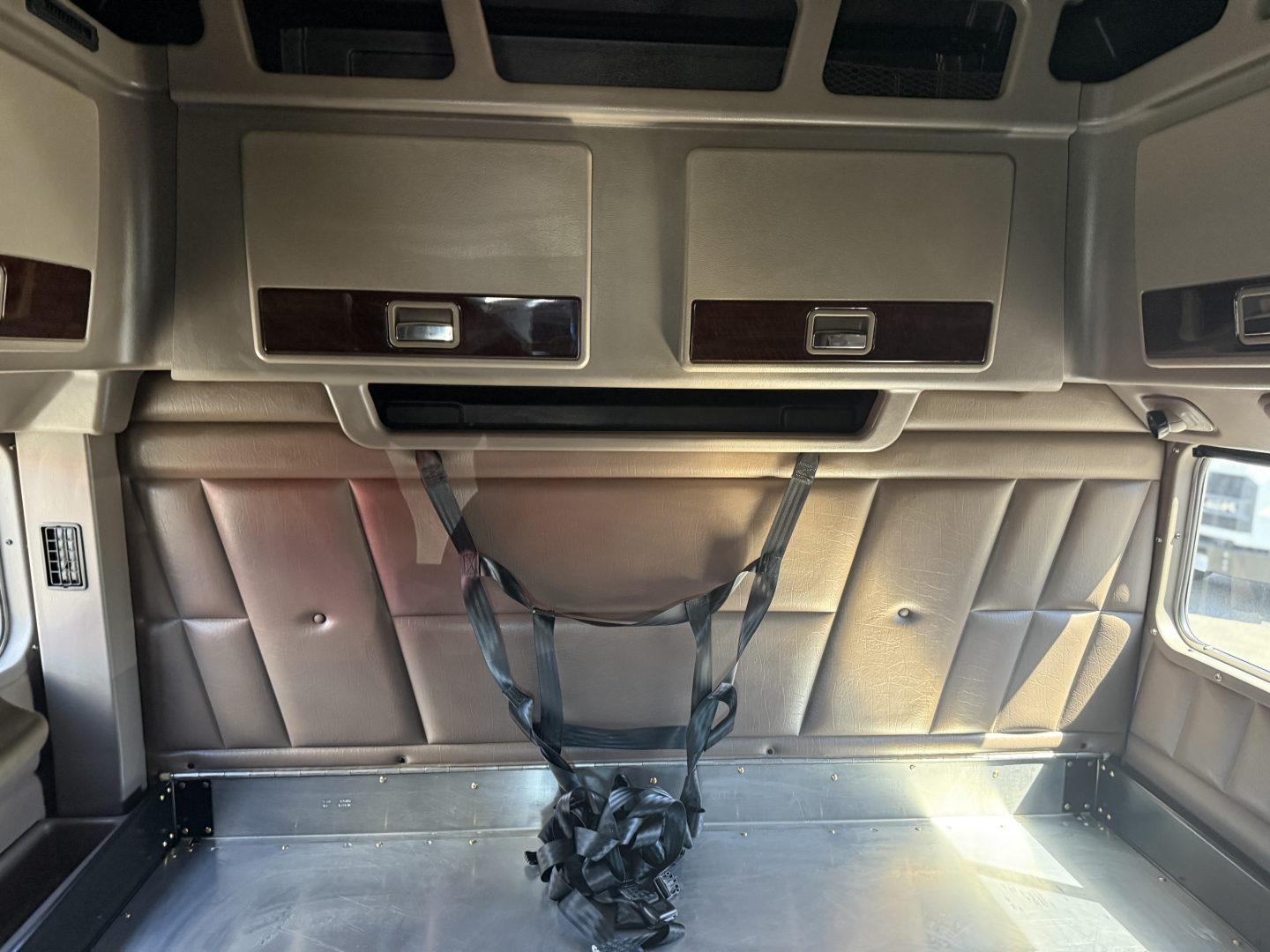 Peterbilt 389 sleeper cab interior cargo area with overhead compartments and fabric panels, tie-down straps in center, light entering from right window.