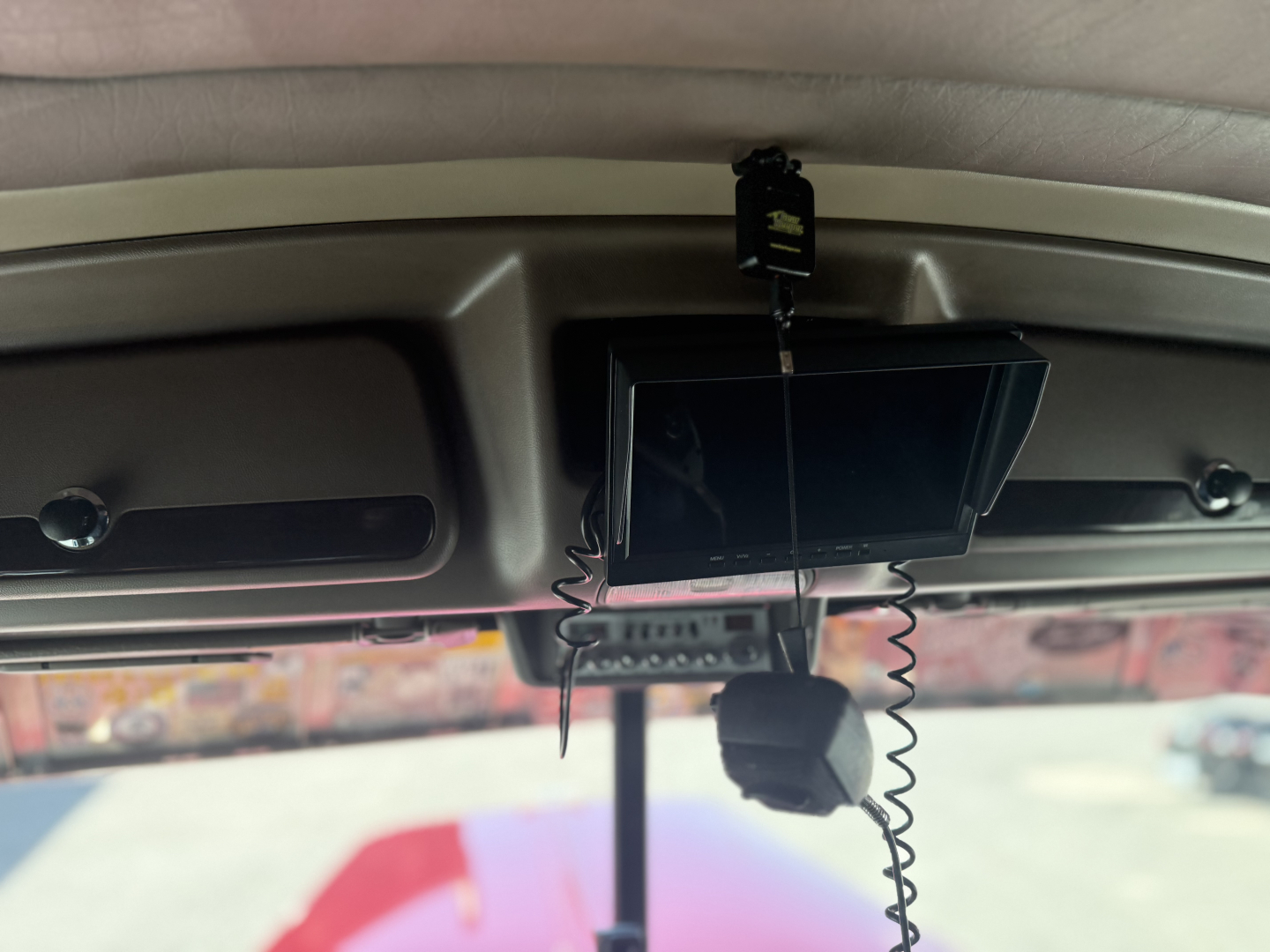 In-cab rearview area with a mounted infotainment screen and a hanging device, visible cords and a small black object; windshield and outside lot visible.