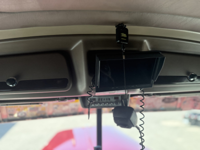 In-cab rearview area with a mounted infotainment screen and a hanging device, visible cords and a small black object; windshield and outside lot visible.