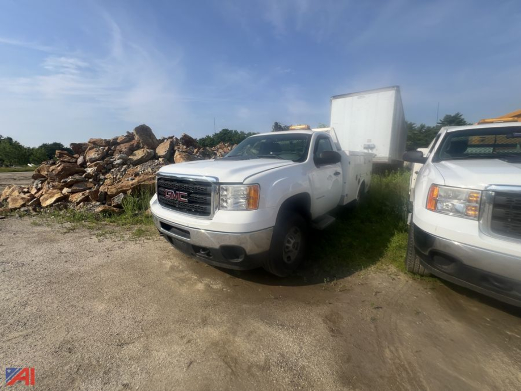 GMC Sierra 2500HD utility truck with crane attachment in a yard, white color, front view, other white utility truck nearby, pile of rocks in background.