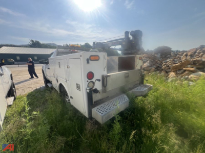 Rear view of a white GMC utility truck with crane equipment, service bed, and metal steps, on a grassy lot.