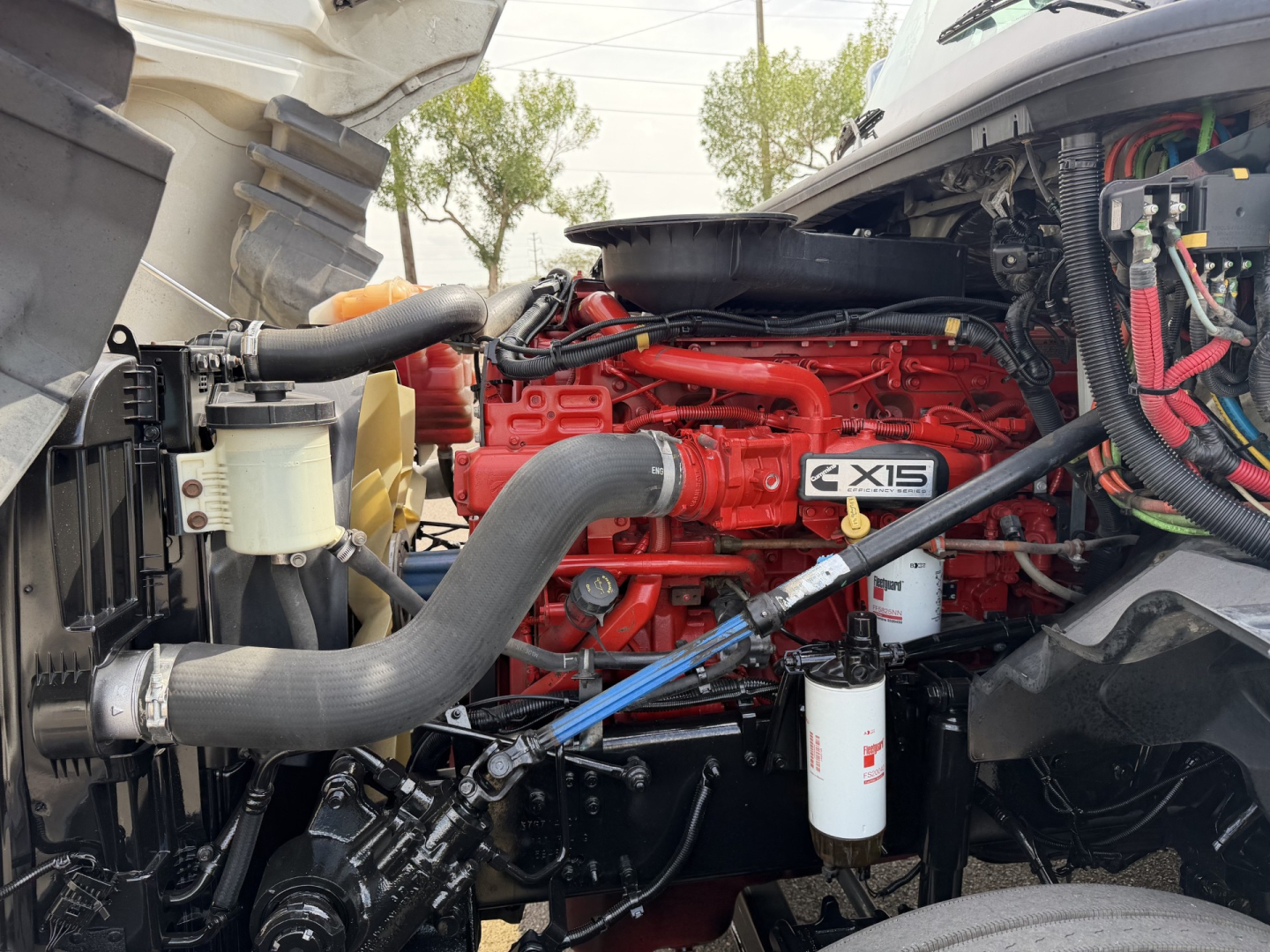 Truck engine bay showing Cummins X15 red engine block and surrounding hoses, coolant reservoir, air intake, and fluid filters.