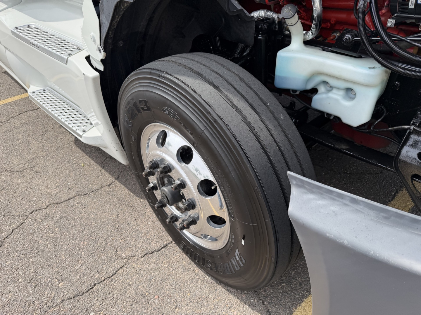 Truck front wheel and engine bay showing dual rear truck tire, chrome hub, white plastic coolant reservoir cap, and part of the engine block.
