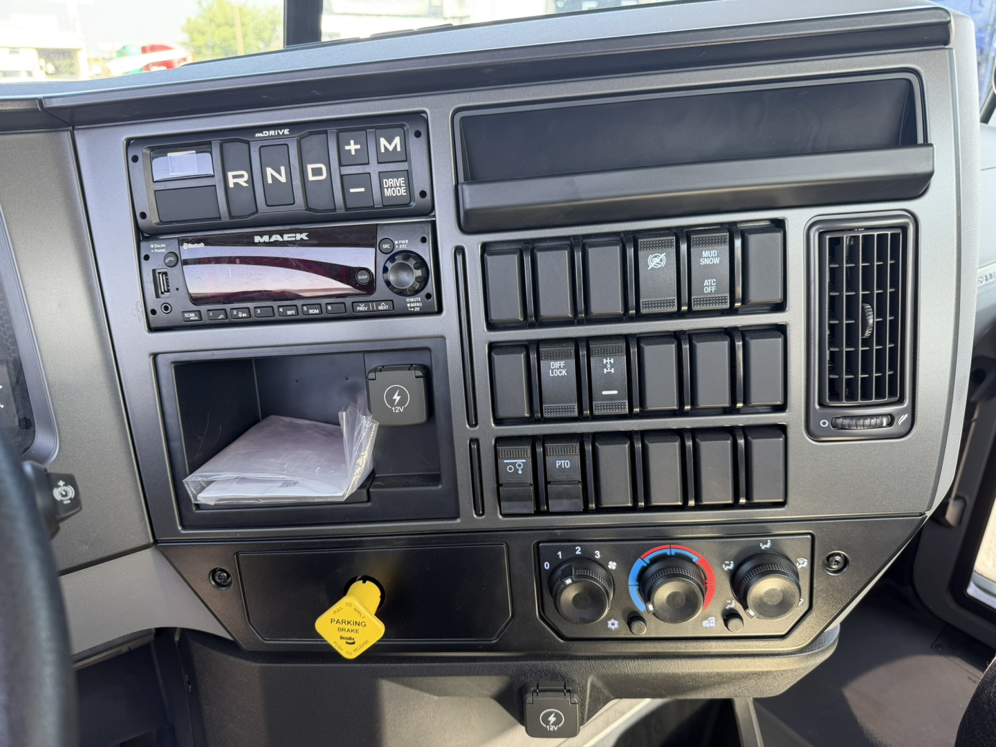 Truck dashboard: gear selector indicators R N D M on left, various switches and a radio in center, row of toggle switches, climate controls with blue/red dials, ignition yellow tag nearby.