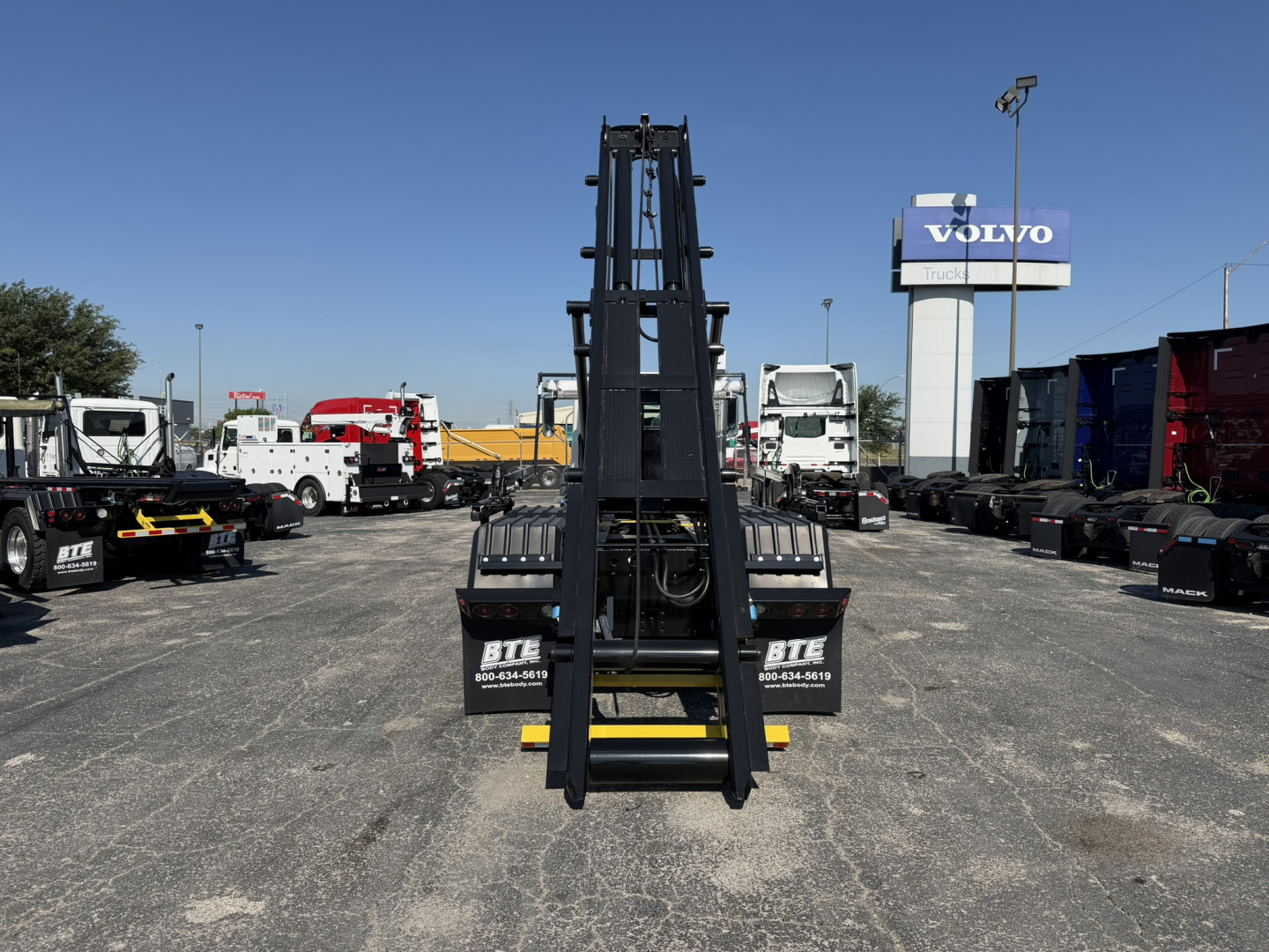 Mack GR64F roll-off truck chassis and hydraulic arm seen from front center in a dealership lot with other trucks in background.