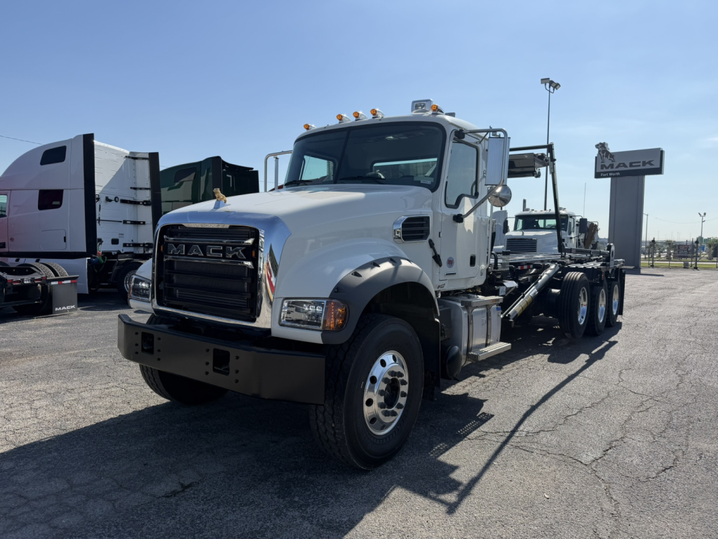 Mack GR64F dump truck chassis in a parking lot showing front cab and wheel, with Mack grille, chrome bumper, and multiple axles in tandem.
