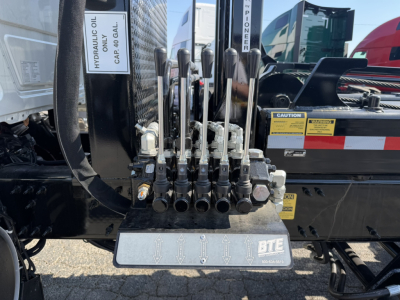 Mack GR64F rack with multiple hydraulic levers and control hoses near a hydraulic oil reservoir and warning labels.
