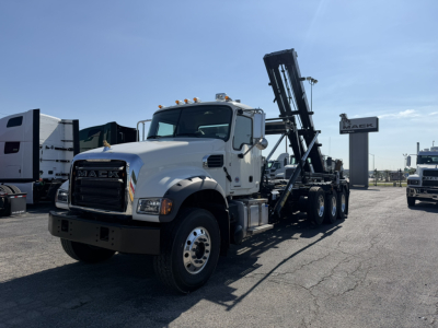 Mack GR64F roll-off truck shown in white, front view with dual rear axles and loading arm raised, Mack logo on grille, clear sky.