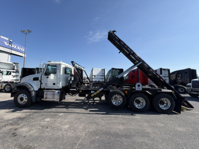 Truck side view showing a Mack GR64F rolloff with tilting dumped bed and multiple axles, white cab, and obvious hydraulic bed lifting mechanism.