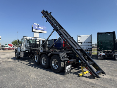 Mack GR64F roll-off truck with loading ramps extended showing stacked black hydraulic booms and triple-axle configuration.