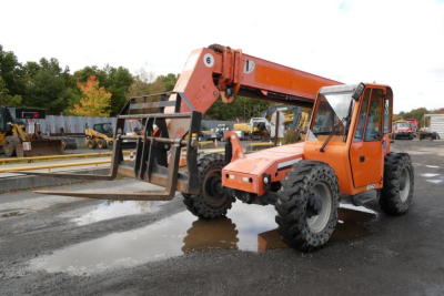 Orange JLG 6042 telehandler in a yard, extended boom with forks, large black tires, cab on right, some puddles in foreground.