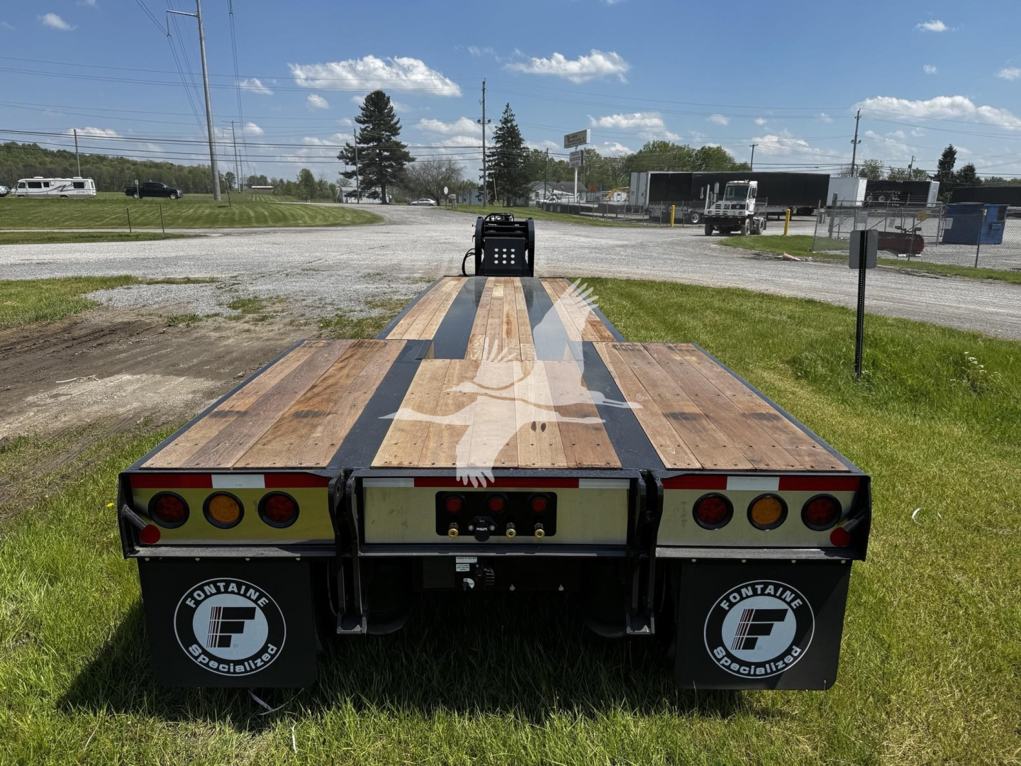 Flatbed trailer rear view showing wooden deck with black stripe runners and round taillights; Fontaine branding on mudflaps; plate area and hitch visible.