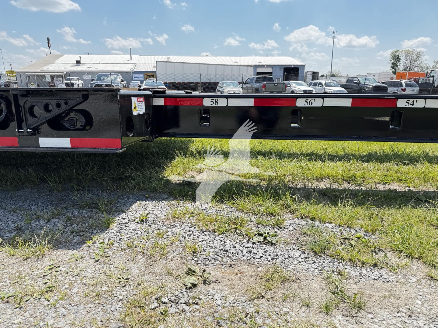 Fontaine lowboy trailer side with numbered ramps '58' '56' '54' and black painted frame; grassy ground in foreground and truck yard in background.