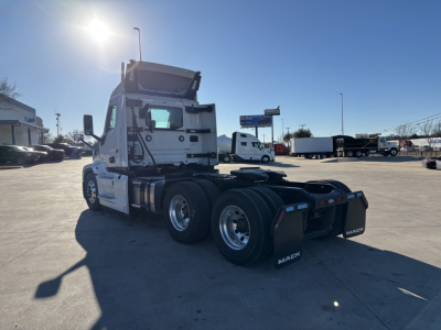Mack PR64T conventional daycab tractor rear view showing triple axles and fifth wheel, with sun glare and dealership lot in background.