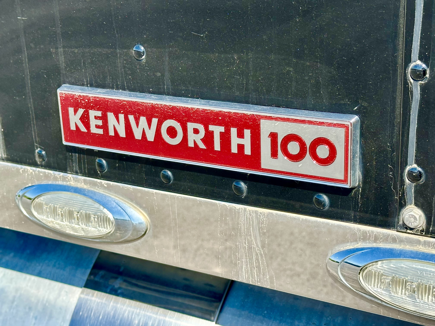 Kenworth 100 emblem on black rear door of a truck with chrome bumper and oval lights visible.