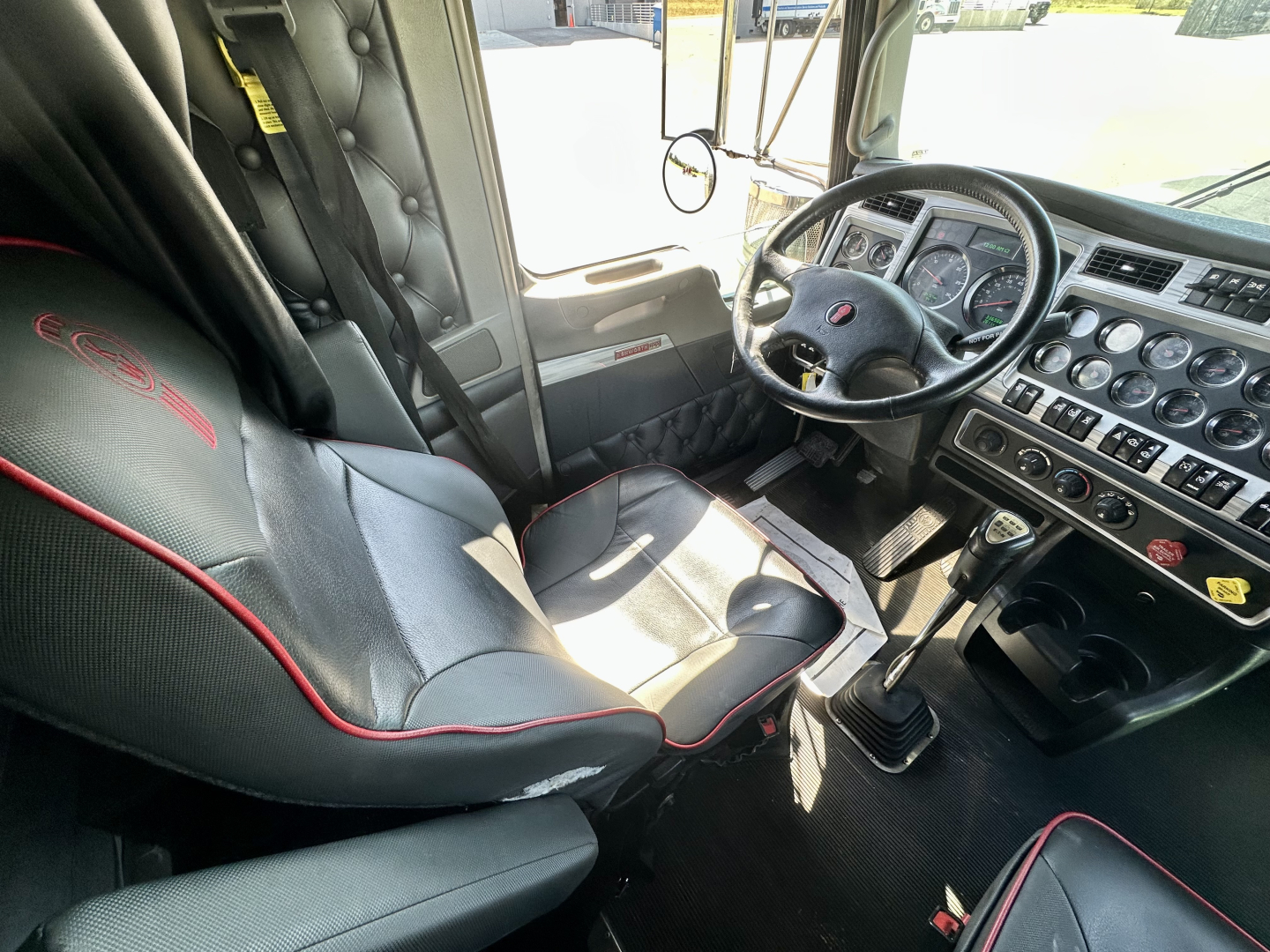 Truck cab interior showing steering wheel, dashboard with numerous gauges, gear shifter, and black seats with red piping.