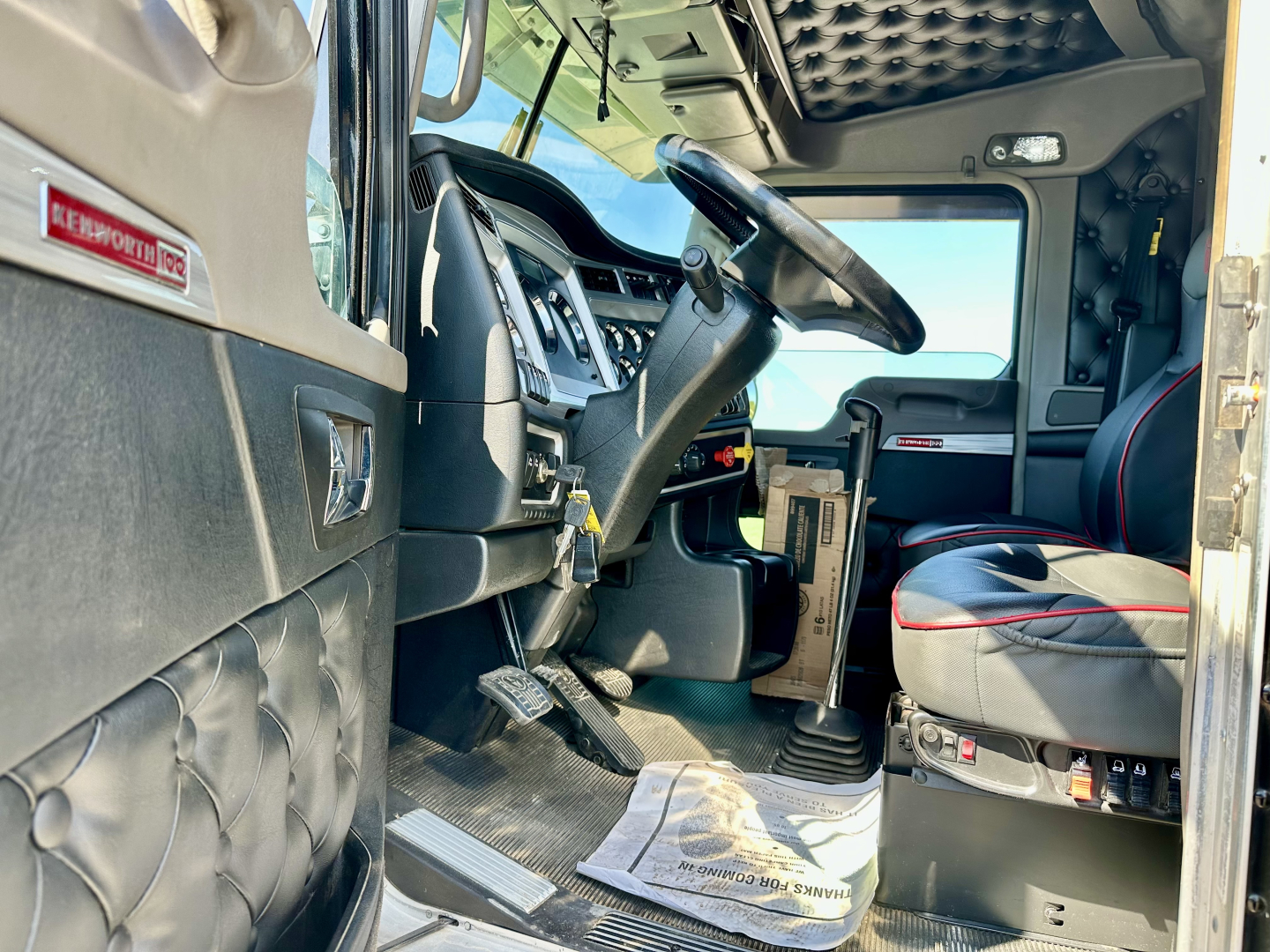 Kenworth W900L truck interior cab view showing steering wheel, dashboard, seat, and pedals with Kenworth logo insignia visible.