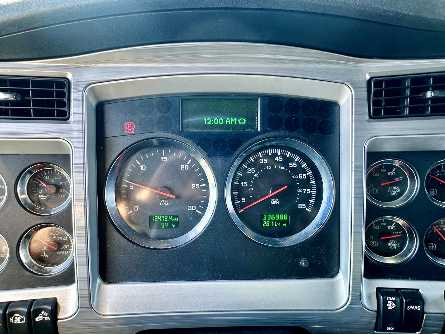 Kenworth W900L dash cluster showing multiple gauges and a green digital clock. Center shows large speedometer and tachometer with odometer. Surrounding gauges with chrome rings and metallic trim.