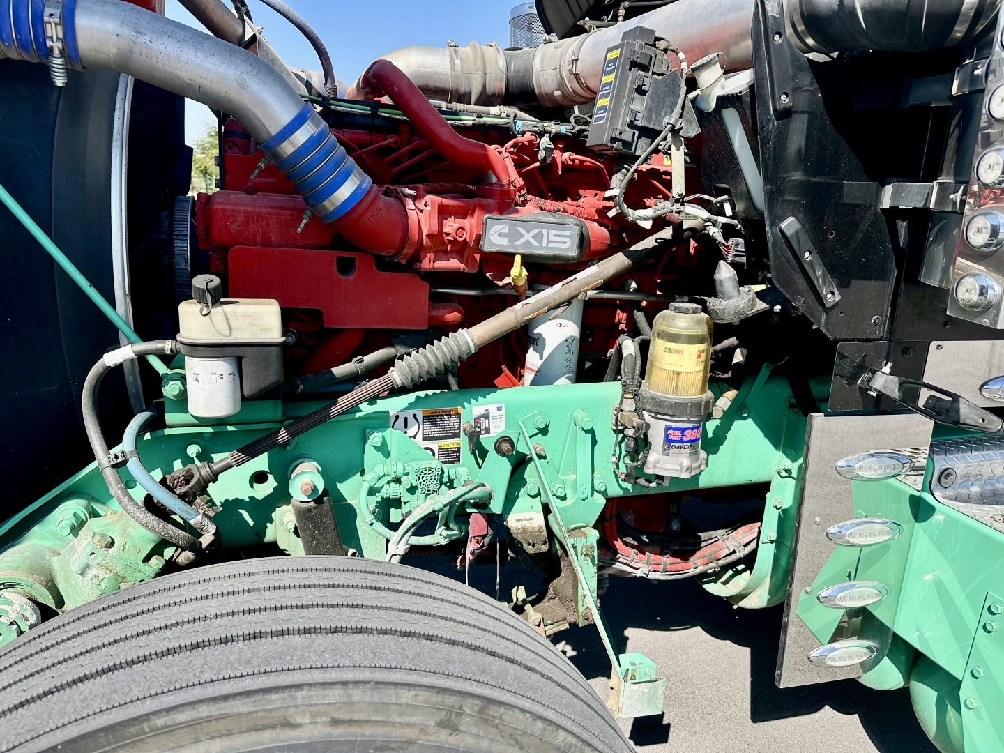 Kenworth W900L engine bay showing 6-cylinder diesel engine colored red with X15 badge, hoses, coolant reservoir, and surrounding green frame; front wheel visible.