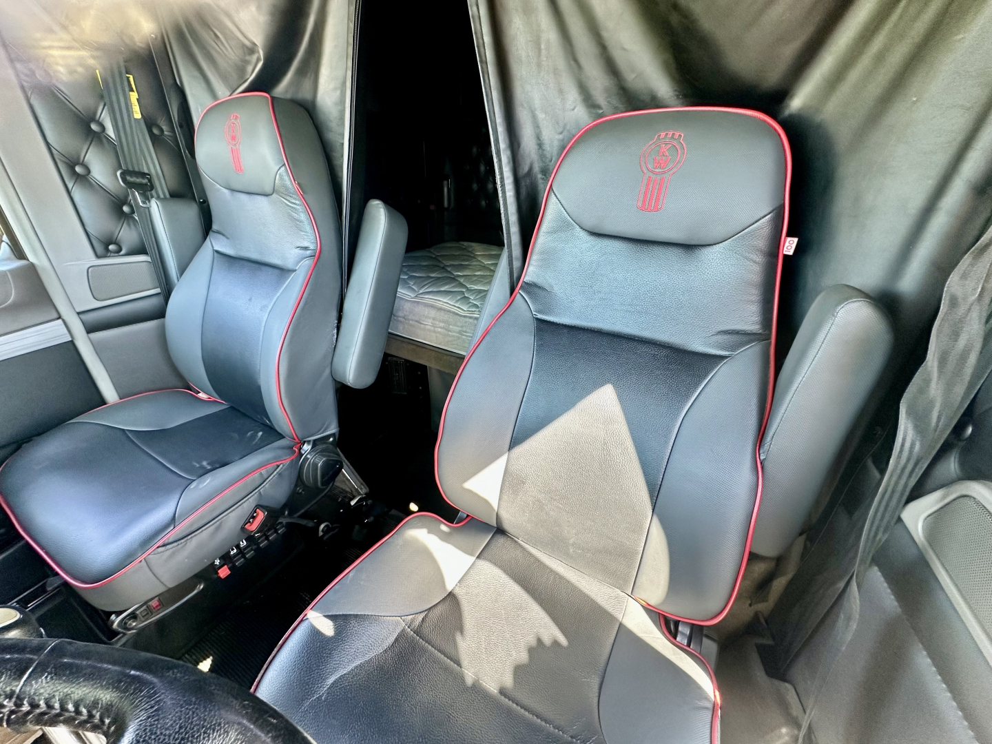 Kenworth W900L truck cab interior showing black leather seats with red piping and embroidered logos. Driver and passenger seats visible with seat adjustments; bunk area behind with curtains.