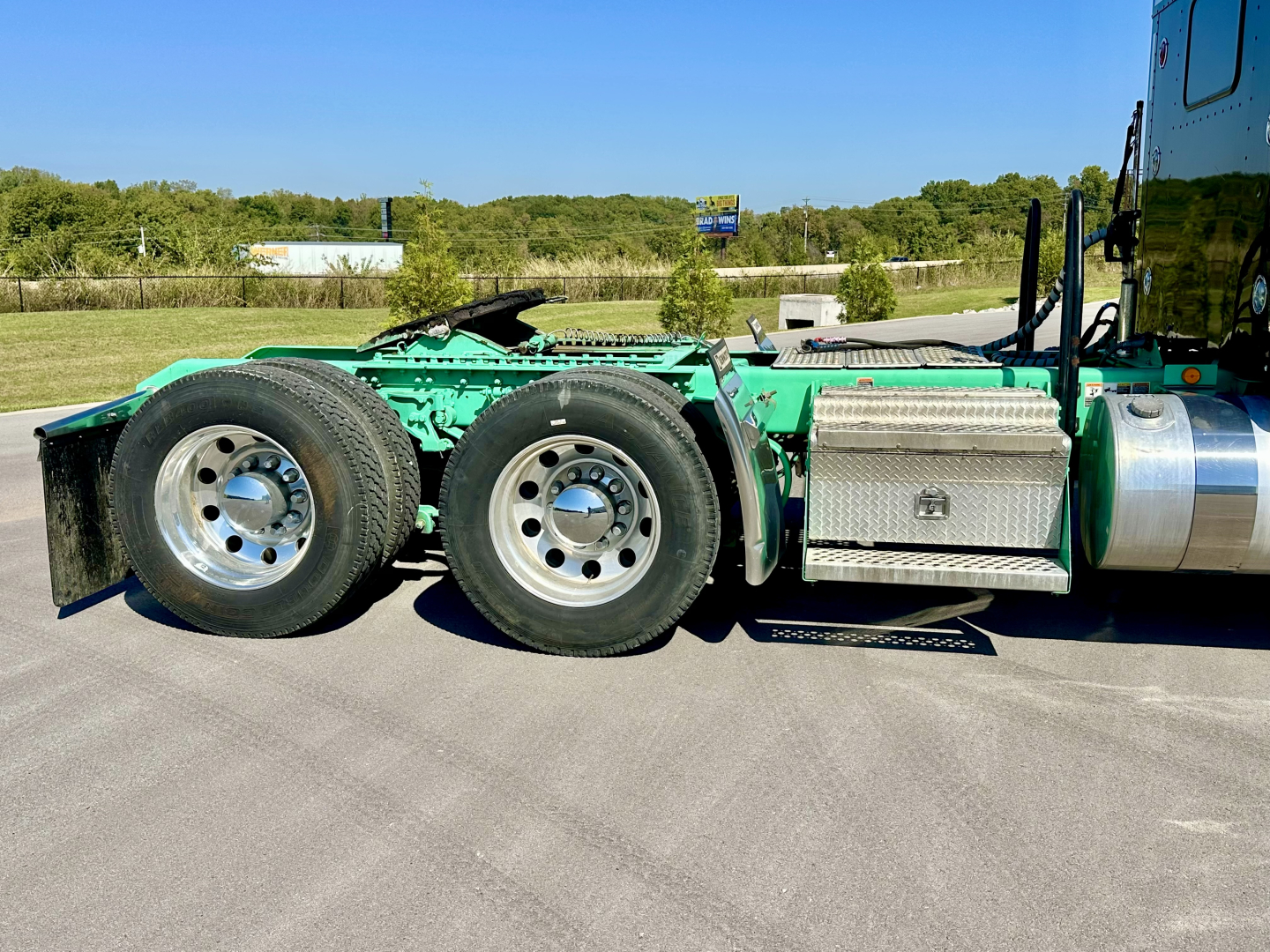 Kenworth W900L truck chassis side view showing rear dual rear axles, green paint, aluminum fuel tank, tool box, and steps.