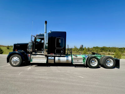 Kenworth W900L truck side view showing engine bay and chrome fuel tanks with black cab and green chassis near-empty lot.