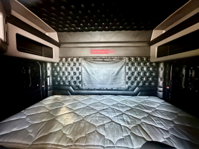 Kenworth W900L sleeper cab interior with quilted headboard and sleeping mattress, storage shelves on sides, overhead lighting panels.