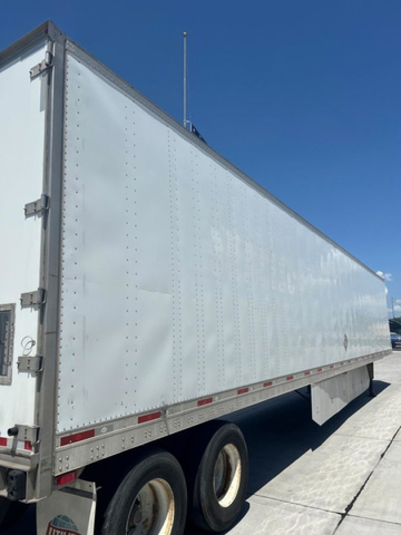 Box trailer side view showing smooth riveted aluminum walls and rear door with hinges; undercarriage with dual rear axles and tires; clear blue sky.