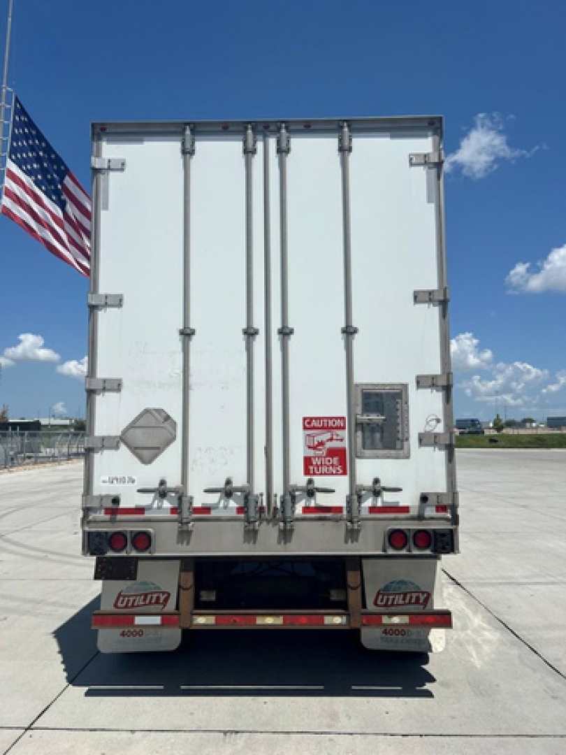 Rear view of a white Utility van trailer with dual rear doors, metal latches, and red warning label, American flag on left, blue sky.
