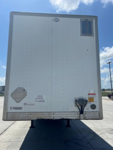 Rear view of a white utility van trailer with riveted door panels, small window/vent near top right, electrical and coupling connections at bottom right, warning label and serial sticker visible.
