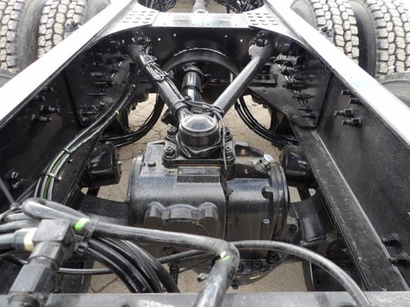 Truck chassis rear view showing driveline with universal joint and drive shaft, axle housing, and multiple hydraulic and electrical cables in engine bay area.