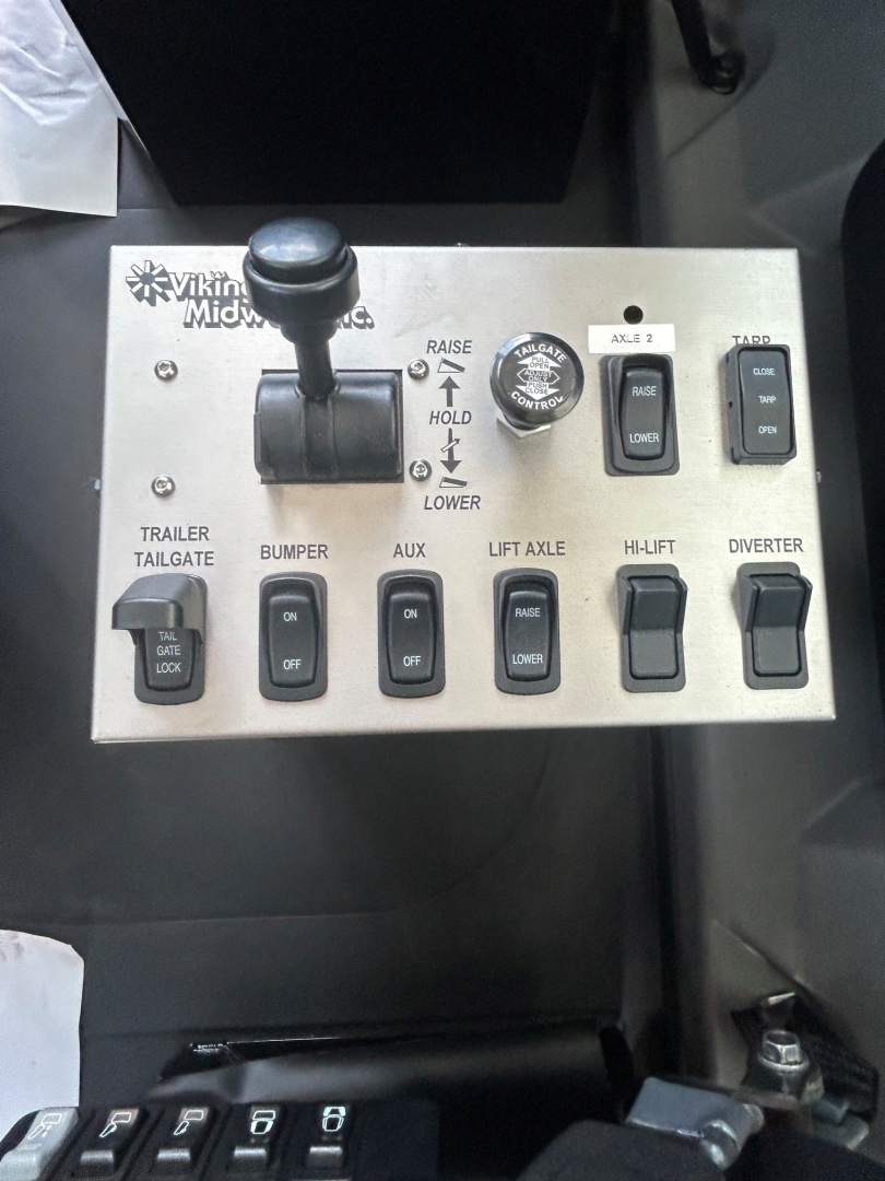 Panel of control switches in Mack GR104B cab showing toggle switches for TRAILER TAILGATE, BUMPER, AUX, LIFT AXLE, HI-LIFT, DIVERTER and a joystick-like lever labeled JOST (RAISE/LOWER) with labels AXLE 2, TARP, etc.
