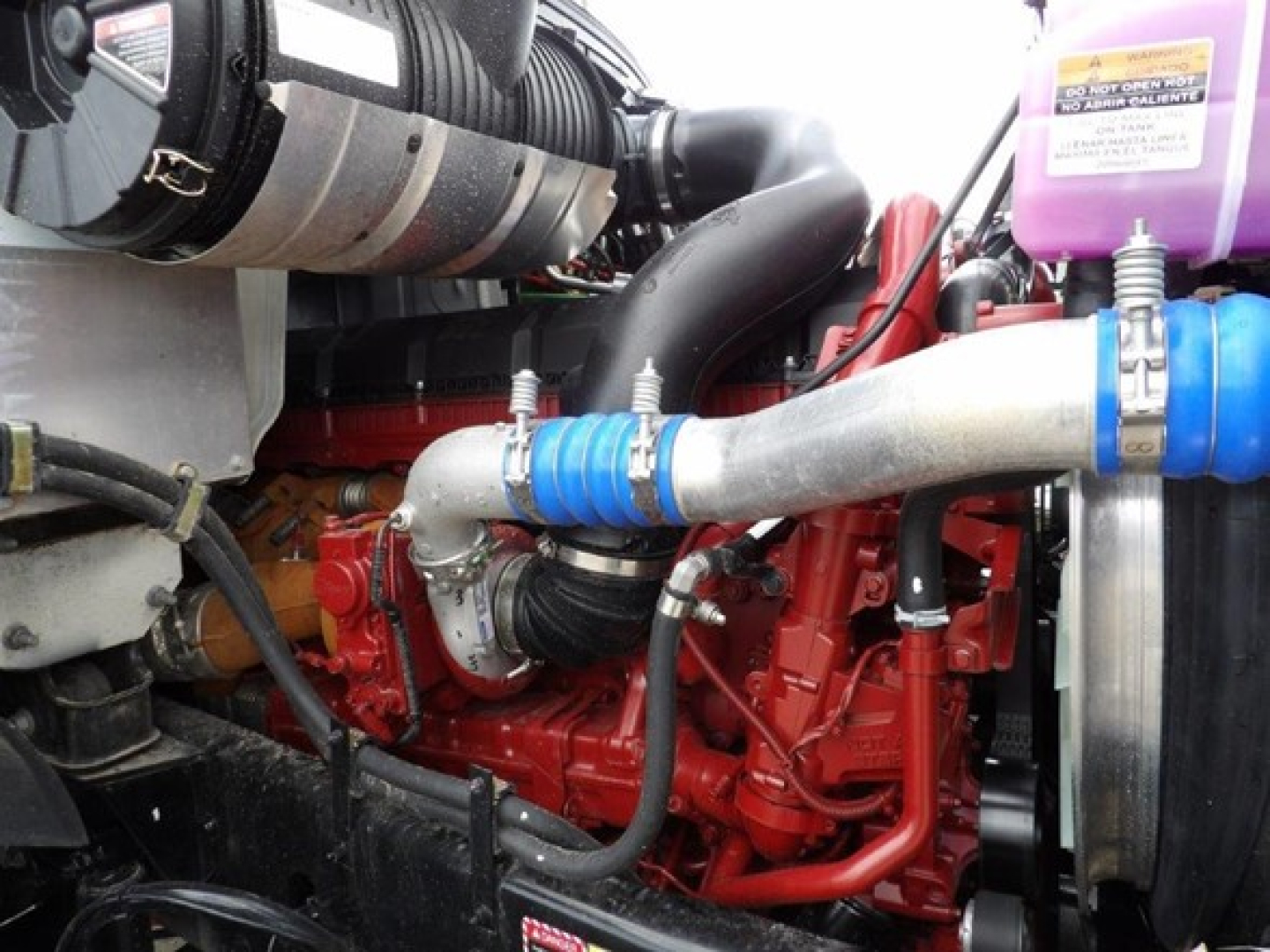Mack GR64B engine bay showing red engine block with blue hose clamps and radiator hose, рядом purple coolant reservoir cap, and intercooler piping.