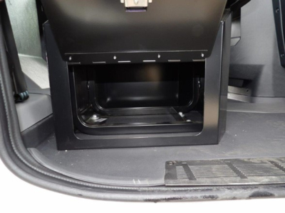 Cab and chassis interior showing storage drawer with empty cavity beneath. Black metal frame, open compartment, entry step lip at bottom, surrounding cabin flooring.