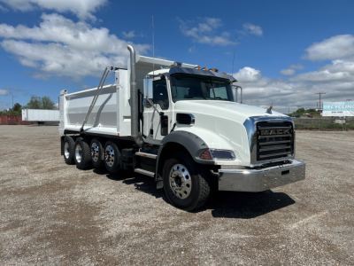 White Mack GR104B dump truck with a large dump bed, chrome front bumper, and multiple rear axles parked on gravel under a blue sky with clouds. Visible text on the truck includes no legible branding in close-up; no other labels readable.