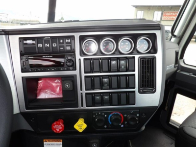 Dash of a 2025 Mack GR64B interior showing RND M shifter, analog gauges, auxiliary switches, climate controls, radio, and warning labels.