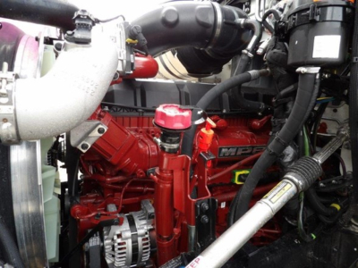 Mack GR64B engine bay showing red diesel engine, radiator hose, coolant reservoir cap, alternator visible, various hoses and wiring.