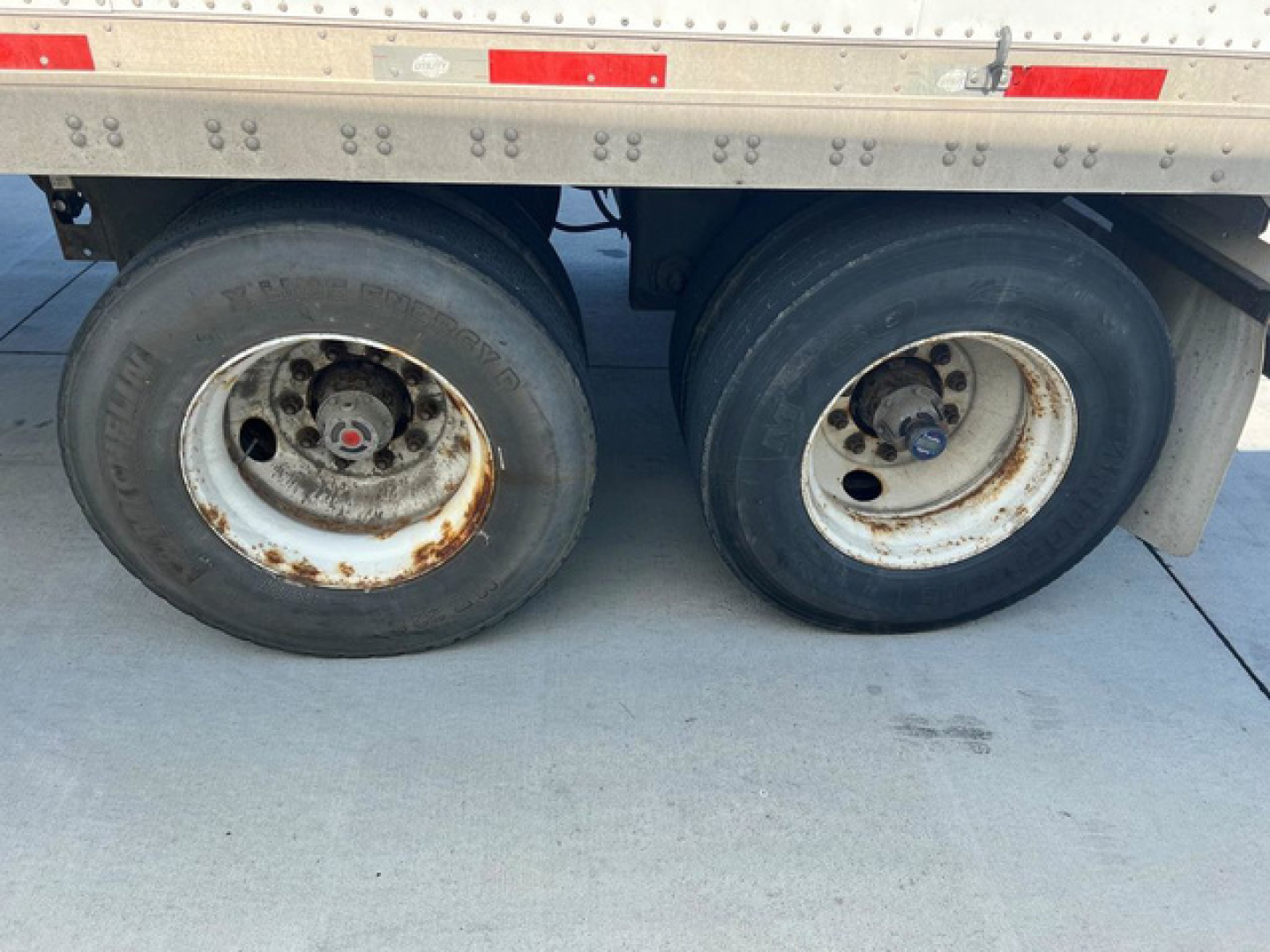 Dual rear trailer tires on a Utility Van showing rusty wheel rims and worn black tires; portion of the trailer side with reflective tape and riveted aluminum side panel.
