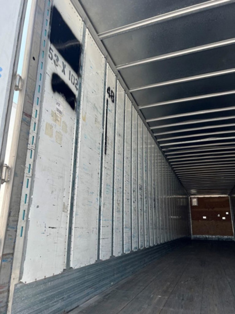 Interior of a trailer with metal side panels and exposed ceiling beams; faint numbers and markings on the walls, long empty cargo area.
