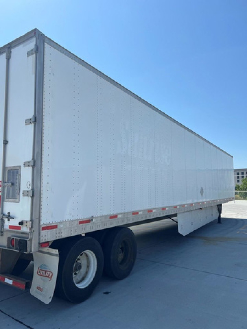 Utility trailer with white box and dual rear wheels. Side shows riveted aluminum panels; rear doors closed. Small Utility logo on mudflap. No legible model text visible.