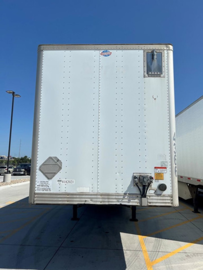 Trailer rear view showing roll-up door, reflective decals, electrical plug, and bumper on a white cargo trailer in a parking lot.