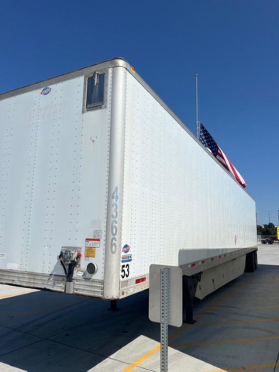 Light-colored utility trailer/van body seen from rear-left, with rear doors and wheel wells, small window near top corner, American flag visible, overhead blue sky.
