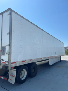 Utility trailer with white box and dual rear wheels. Side shows riveted aluminum panels; rear doors closed. Small Utility logo on mudflap. No legible model text visible.