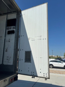 Rear of a utility van trailer with an open rear door showing an empty interior and a ventilated panel on the side, clear blue sky.