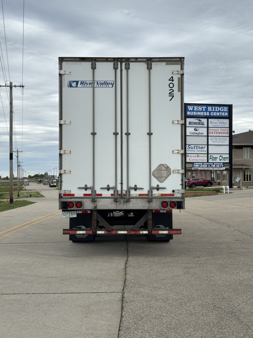 Rear view of a white Utility trailer with double doors. Visible text includes River Valley logo on left door and the number 4027 on right edge. Street signs in distance, overcast sky.