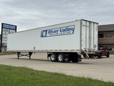 Trailer: River Valley Trailer Leasing logo on side of long white utility trailer with dual rear doors and a red pickup at front; parked on paved lot.