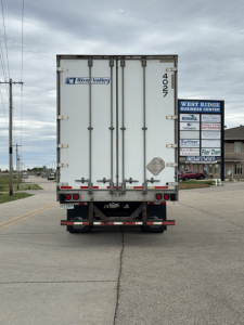 Rear view of a white Utility trailer with double doors. Visible text includes River Valley logo on left door and the number 4027 on right edge. Street signs in distance, overcast sky.