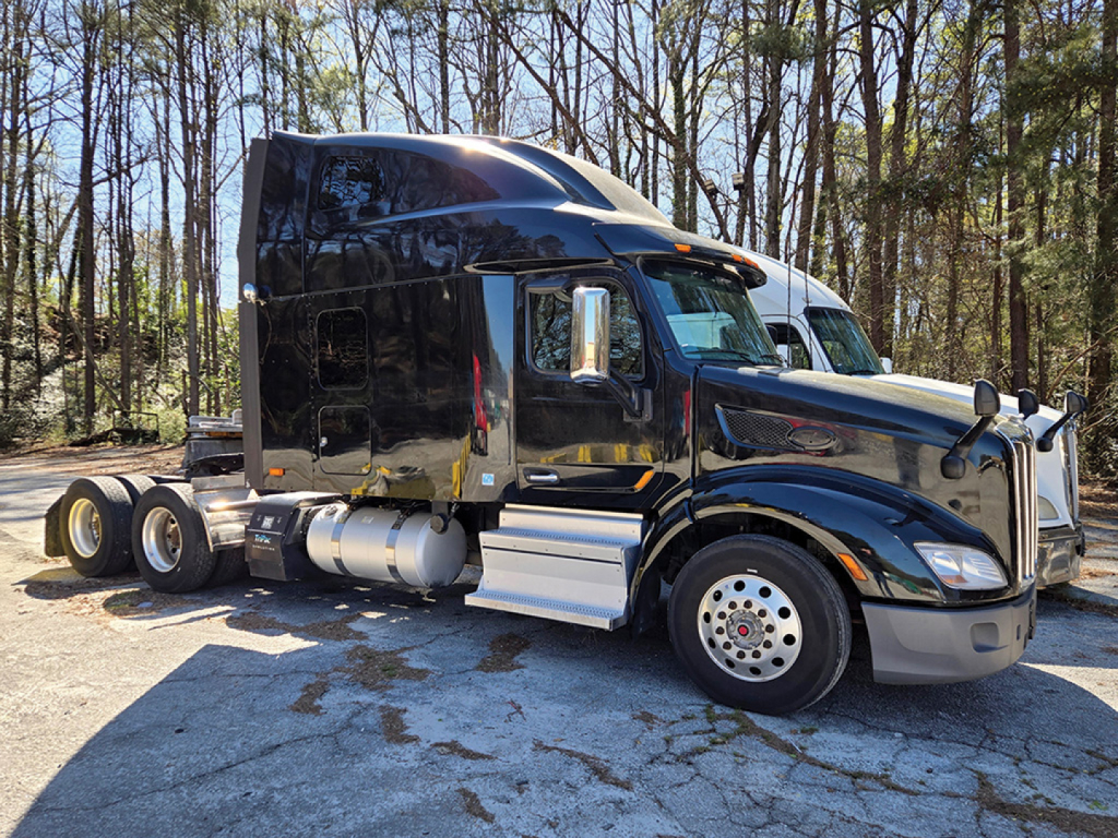 Peterbilt 579 truck in a parking lot, black cab, sleeper, diesel fuel tanks visible, side view showing wheels and windows.