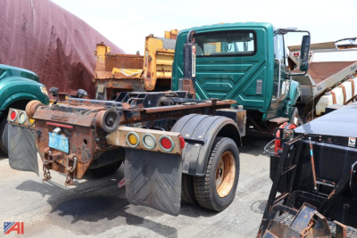Truck chassis with hooklift aft view showing rusty rear frame and dual rear wheels, International TerraStar truck in green cab, other equipment in background.